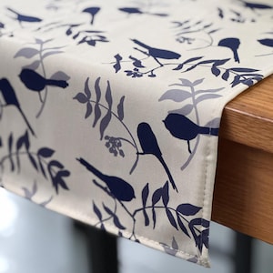 May include: A rectangular table runner featuring a cream-colored background with a repeating pattern of dark blue birds perched on branches with gray and blue leaves. The fabric has a finished hem along the edge and drapes over a wooden surface.