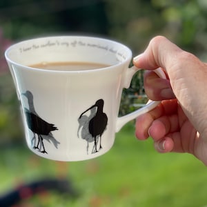 May include: A white ceramic teacup with a black silhouette of three birds. The cup has a quote on the rim that reads "I have the ocean's very salt, the mountain's cloud and mist".