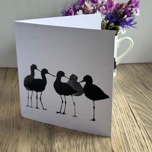 Könnte beinhalten: Eine weiße, gefaltete Grußkarte mit einer schwarzen Tuschezeichnung von vier Watvögeln, die in einer Reihe stehen. Die Vögel sind als Silhouetten mit feinen punktierten Texturdetails auf ihren Körpern dargestellt und durch ihre langen, dünnen Schnäbel verbunden.