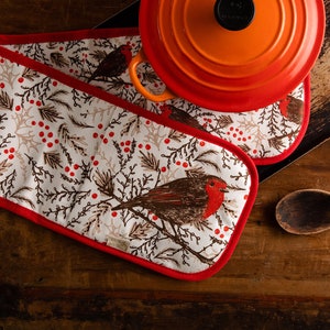 May include: Two red and white oven mitts with a robin and holly pattern. The mitts are on a wooden surface with a red and orange pot and a wooden spoon.