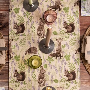 May include: A beige table runner with a woodland pattern featuring foxes, squirrels, hedgehogs, and bluebells. The runner is decorated with candles and candle holders.