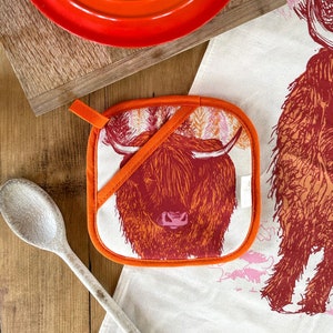 Puede incluir: Un agarrador de olla naranja y rojo con un diseño de vaca de las Tierras Altas. La vaca es marrón y tiene la cara blanca. El agarrador de olla tiene un lazo para colgar.