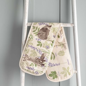May include: A pair of beige oven mitts with a floral and woodland creature design. The mitts feature a hedgehog, a fox, and various plants with their names printed on the fabric. The mitts are hanging on a white wooden ladder.