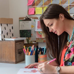 Puede incluir: Una mujer con una camisa de estampado floral est&aacute; sentada en un escritorio y coloreando un dibujo con marcadores rosas. Lleva un collar de cuentas turquesa. Hay marcadores de colores en dos vasos sobre el escritorio.