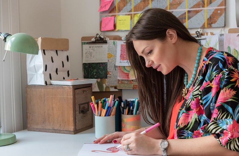 Puede incluir: Una mujer con una camisa de estampado floral est&aacute; sentada en un escritorio y coloreando un dibujo con marcadores rosas. Lleva un collar de cuentas turquesa. Hay marcadores de colores en dos vasos sobre el escritorio.