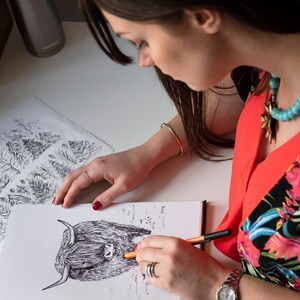 Puede incluir: Una mujer est&aacute; dibujando una ilustraci&oacute;n en blanco y negro de una vaca de las tierras altas con un bol&iacute;grafo y un l&aacute;piz naranja. El dibujo est&aacute; en una hoja de papel blanca. La mujer lleva una camisa roja con estampado floral y un collar turquesa.