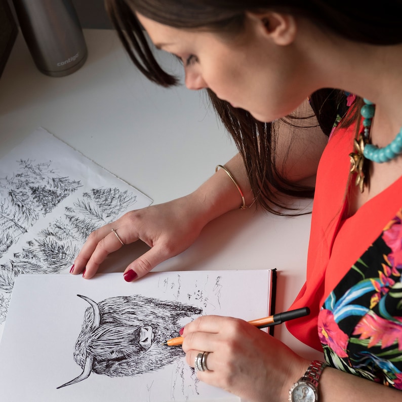 Puede incluir: Una mujer est&aacute; dibujando una ilustraci&oacute;n en blanco y negro de una vaca de las tierras altas con un bol&iacute;grafo y un l&aacute;piz naranja. El dibujo est&aacute; en una hoja de papel blanca. La mujer lleva una camisa roja con estampado floral y un collar turquesa.
