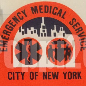 May include: A red circular sticker with the text "Emergency Medical Service City of New York". The sticker features a black and white illustration of a city skyline, a medical symbol, and a group of people.