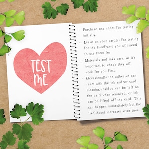 May include: Open notebook with a red heart that says "TEST ME" in white. The notebook is open to a page with instructions. Green leaves and sprigs are arranged around the notebook on a brown surface.