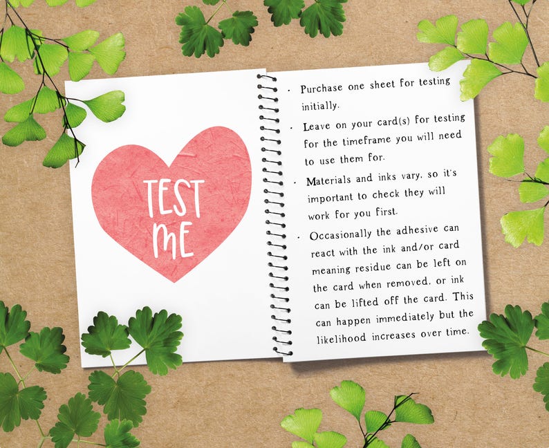 May include: Open notebook with a red heart that says "TEST ME" in white. The notebook is open to a page with instructions. Green leaves and sprigs are arranged around the notebook on a brown surface.