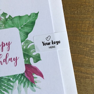 May include: A white greeting card with a watercolor design of green tropical leaves and pink accents. The card has the words "Happy Birthday" in pink script. A clear sticker with a heart and the words "Your Logo HERE" is on the card.