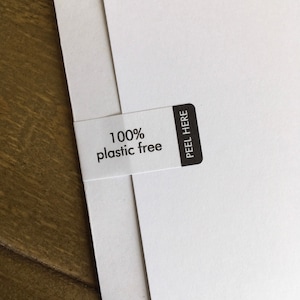 May include: Close-up of white paper sheets with a white label that reads "100% plastic free." The label has a black rectangle with the words "PEEL HERE." The background is a wooden surface.