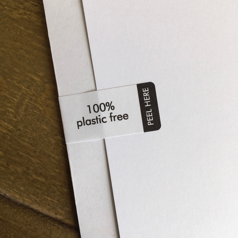 May include: Close-up of white paper sheets with a white label that reads "100% plastic free." The label has a black rectangle with the words "PEEL HERE." The background is a wooden surface.
