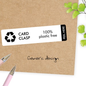 May include: A rectangular white label with the text "CARD CLASP" and a recycling symbol, along with "100% plastic free". The label is on a brown paper background. The words "PEEL HERE" are on the right side of the label. Two pink pens are in the lower left corner.