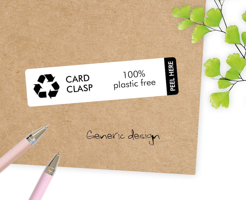 May include: A rectangular white label with the text "CARD CLASP" and a recycling symbol, along with "100% plastic free". The label is on a brown paper background. The words "PEEL HERE" are on the right side of the label. Two pink pens are in the lower left corner.