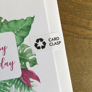 May include: White card with a watercolor illustration of green tropical leaves and a pink flower. A white label with the text "CARD CLASP" and a recycling symbol is attached to the card.