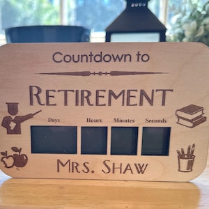 May include: Wooden retirement countdown sign. The sign reads "Countdown to Retirement" and "Mrs. Shaw". It features spaces for days, hours, minutes, and seconds. Includes teacher-themed graphics such as apples, books, and pencils.