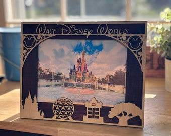 Op de afbeelding: Een houten fotolijst met een uitgesneden ontwerp met Mickey Mouse-oren, een kasteel en een bol. De lijst heeft een donkerbruine rand en de tekst "Walt Disney World" is in wit bovenaan gedrukt. De lijst toont een foto van Assepoesterkasteel in Walt Disney World.