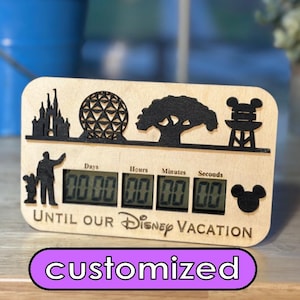 May include: A wooden countdown clock featuring black and white silhouettes of Disney landmarks, including Cinderella's Castle, Spaceship Earth, the Tree of Life, and a Mickey Mouse icon. The clock displays days, hours, minutes, and seconds. The text "Until Our Disney Vacation" is printed below the clock. The word "customized" is printed in purple on a white background below the clock.