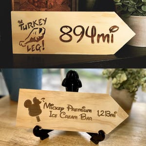 May include: Wooden arrow-shaped signs with engraved text and images. One sign reads "Turkey Leg!" and "894 mi." The other sign says "Mickey Premium Ice Cream Bar" and "1,213 mi."
