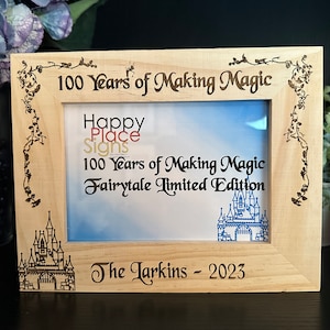 May include: A wooden picture frame with a light brown finish. The frame is engraved with the text "100 Years of Making Magic", "Happy Place Signs", "100 Years of Making Magic Fairytale Limited Edition", and "The Larkins - 2023".