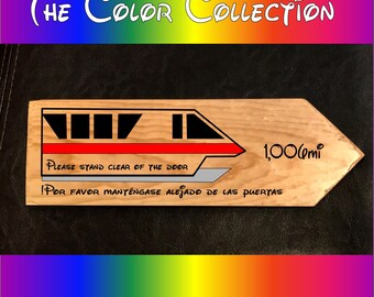 May include: Wooden sign with a painted image of a monorail, the text "PLEASE STAND CLEAR OF THE DOOR" and "!POR FAVOR MANTÉNGASE ALEJADO DE LAS PUERTAS" and the text "1,000mi".
