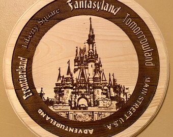 May include: A round wooden sign with a brown border and a light wood background. The sign features a black and white image of Cinderella's Castle at Walt Disney World. The border of the sign has the names of the different lands at Walt Disney World: Adventureland, Liberty Square, Frontierland, Fantasyland, Tomorrowland, Main Street U.S.A.