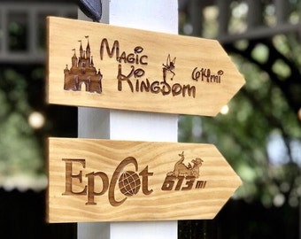 May include: Two wooden signs with arrows pointing in opposite directions. The top sign says "Magic Kingdom 0.4 mi" with a silhouette of Cinderella's Castle and a fairy. The bottom sign says "Epcot 6.73 mi" with a silhouette of the Epcot ball and a dragon.