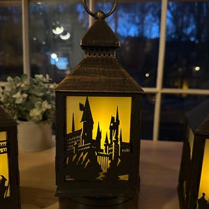 May include: Black metal lantern with a castle silhouette design, illuminated by a warm yellow light. The lantern has a handle at the top and sits on a round base. The design is reminiscent of a famous wizarding school.