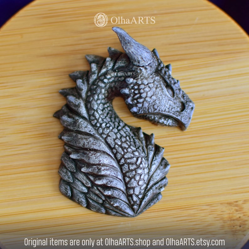 May include: A silver-colored dragon head sculpture with detailed scales and a textured finish. The sculpture is on a wooden background.