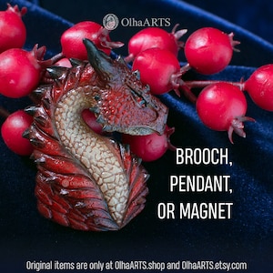 Può includere: Una scultura di testa di drago rossa realizzata in argilla polimerica. Il drago ha occhi neri e una pancia bianca. La scultura è disponibile come spilla, ciondolo o calamita. Gli articoli originali sono disponibili solo su OlhaARTS.shop e OlhaARTS.etsy.com