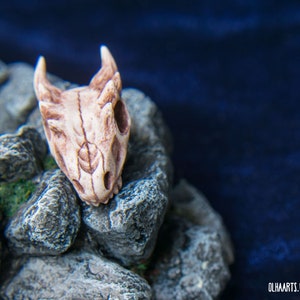 May include: A small, white, detailed dragon skull charm with a textured surface. The skull is resting on a pile of gray rocks.
