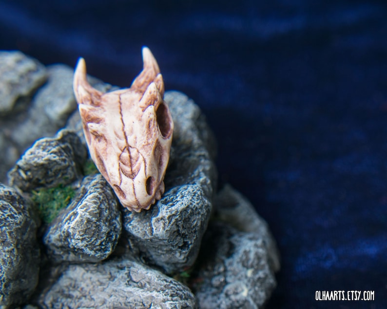 May include: A small, white, detailed dragon skull charm with a textured surface. The skull is resting on a pile of gray rocks.