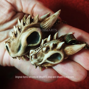 May include: Two small, detailed, bone-colored dragon skull pendants with a textured surface. The pendants are on a chain and are held in a hand. The text "OlhaARTS" is visible in the image.