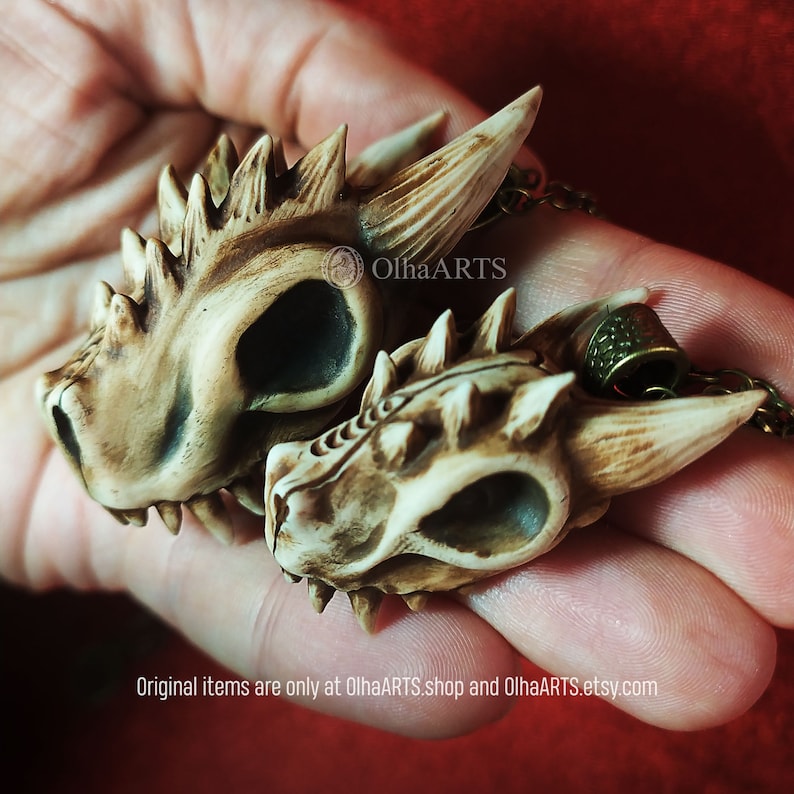 May include: Two small, detailed, bone-colored dragon skull pendants with a textured surface. The pendants are on a chain and are held in a hand. The text "OlhaARTS" is visible in the image.