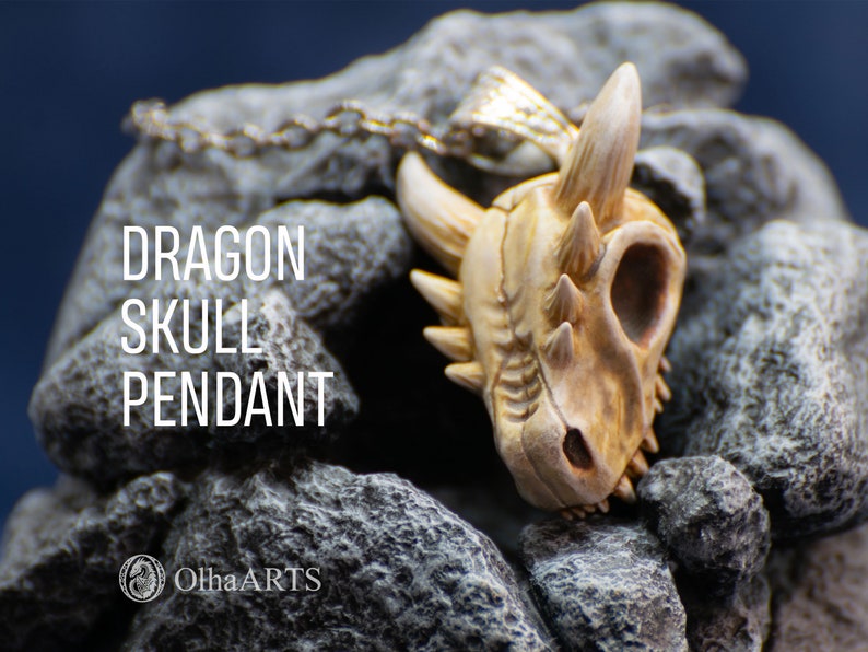May include: A detailed dragon skull pendant made of white and beige polymer clay. The pendant is hanging from a silver chain and is resting on a bed of gray rocks. The text "DRAGON SKULL PENDANT" is visible in the image.