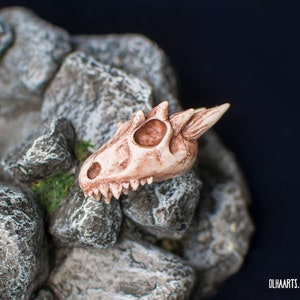 May include: A small, detailed, beige-colored dragon skull with a pointed horn on top, resting on a bed of gray rocks.