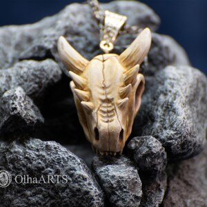 May include: A white ceramic dragon skull pendant with a silver chain. The pendant is detailed with sharp teeth and horns, and is set against a backdrop of gray rocks.