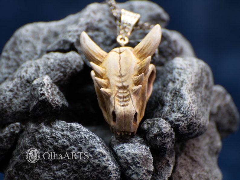 May include: A white ceramic dragon skull pendant with a silver chain. The pendant is detailed with sharp teeth and horns, and is set against a backdrop of gray rocks.