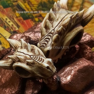 May include: Two detailed, white dragon skull figurines with brown accents. The skulls are resting on a copper-colored surface. Original items are only at OlhaARTS shop and OlhaARTS.etsy.com.