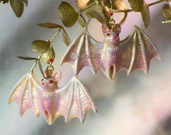May include: A pair of iridescent pink bat earrings with red eyes. The bats are hanging from gold hoops with red beads.