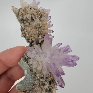 May include: A cluster of amethyst crystals on a matrix of rock. The crystals are a light purple color and have a pointed shape. The matrix is a light brown color and has a rough texture.