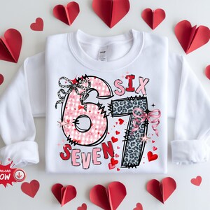 Puede incluir: Sudadera blanca de manga larga con un diseño gráfico que presenta los números "6" y "7" en cuadros rosas y estampado de leopardo, con la palabra "SEVEN" debajo. Corazones de papel rojo rodean la camisa.