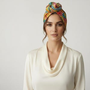 Può includere: Un turbante colorato con un motivo floreale tropicale nei toni del rosso, giallo, verde e blu. Il turbante è annodato sulla sommità e indossato sulla testa. La persona indossa una camicetta color crema con scollatura a cascata.