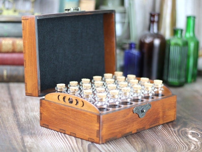 May include: A wooden box with a hinged lid, containing small glass bottles with cork stoppers. The box is stained a warm brown color and has a black felt interior. The bottles are arranged in rows. The box is open, revealing the contents.