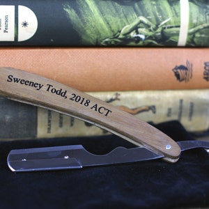 Peut inclure: Un rasoir droit avec un manche en bois, grav&eacute; de "Sweeney Todd, 2018 ACT", est pos&eacute; sur une surface noire. La lame du rasoir est argent&eacute;e. En arri&egrave;re-plan, une pile de livres anciens, dont "Escape Stories".