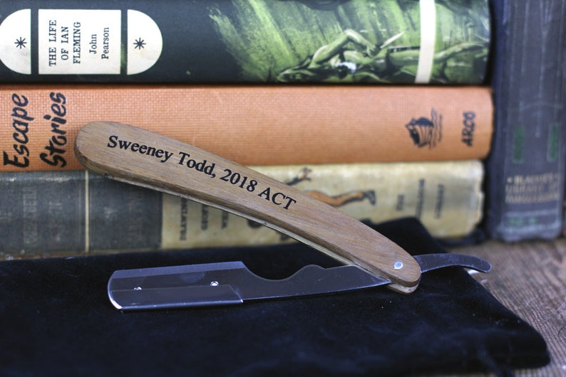 Peut inclure: Un rasoir droit avec un manche en bois, grav&eacute; de "Sweeney Todd, 2018 ACT", est pos&eacute; sur une surface noire. La lame du rasoir est argent&eacute;e. En arri&egrave;re-plan, une pile de livres anciens, dont "Escape Stories".