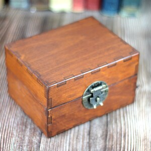 May include: A rectangular wooden box with a hinged lid and a metal clasp. The box is stained a warm brown color, with visible wood grain. The metal clasp is a tarnished brass color. The box is likely used for storage or display.