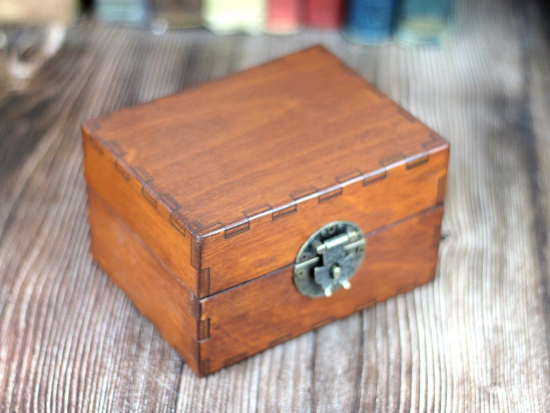 May include: A rectangular wooden box with a hinged lid and a metal clasp. The box is stained a warm brown color, with visible wood grain. The metal clasp is a tarnished brass color. The box is likely used for storage or display.