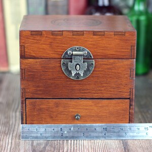 May include: A wooden box with a drawer and a hinged lid, featuring a decorative metal clasp. The box is stained a warm brown color. The box measures approximately 6 inches wide.